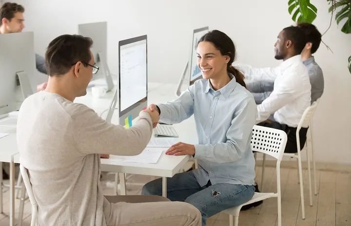 Two colleagues smiling and shaking hands across a desk in a modern office with computers and other coworkers in the background.