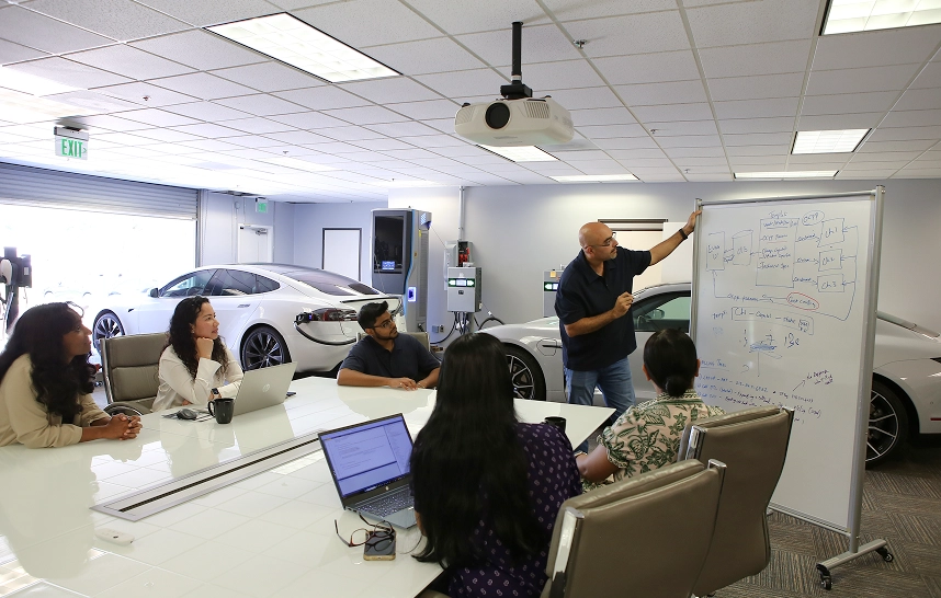 Man presenting to a group in a conference room with a whiteboard and electric cars in the background.