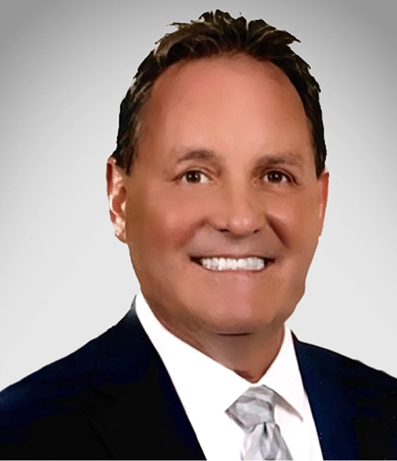 Smiling man in a dark suit and light gray patterned tie against a plain gray background.