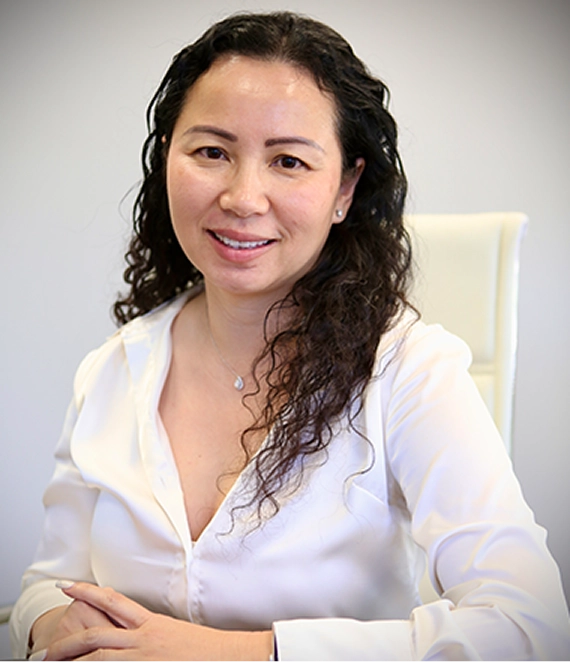 Smiling woman with curly dark hair wearing a white blouse sitting on a white chair.