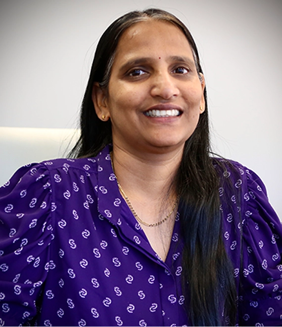 Smiling woman with long dark hair wearing a purple patterned shirt and gold jewelry.