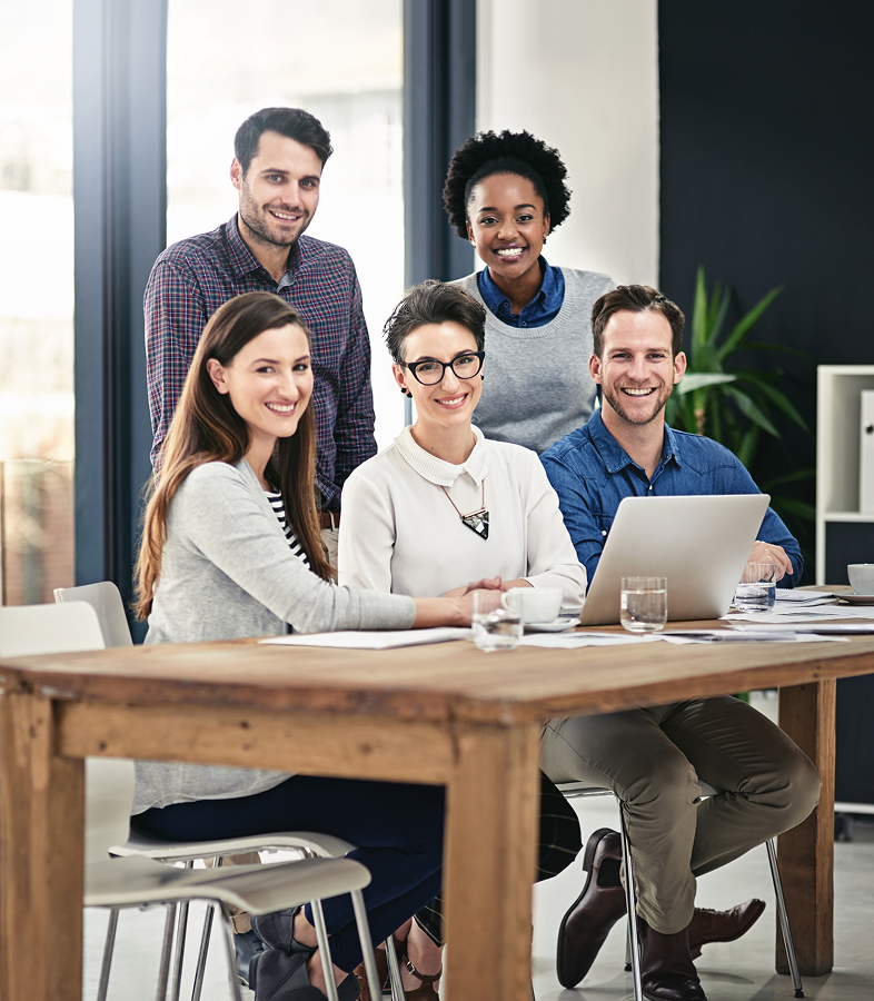Five diverse professionals smiling around a wooden table with a laptop and glasses in a modern office.