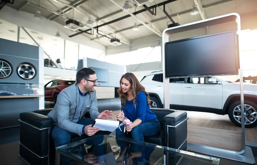 Man in a gray blazer discusses paperwork with a woman in a blue shirt at a car dealership.