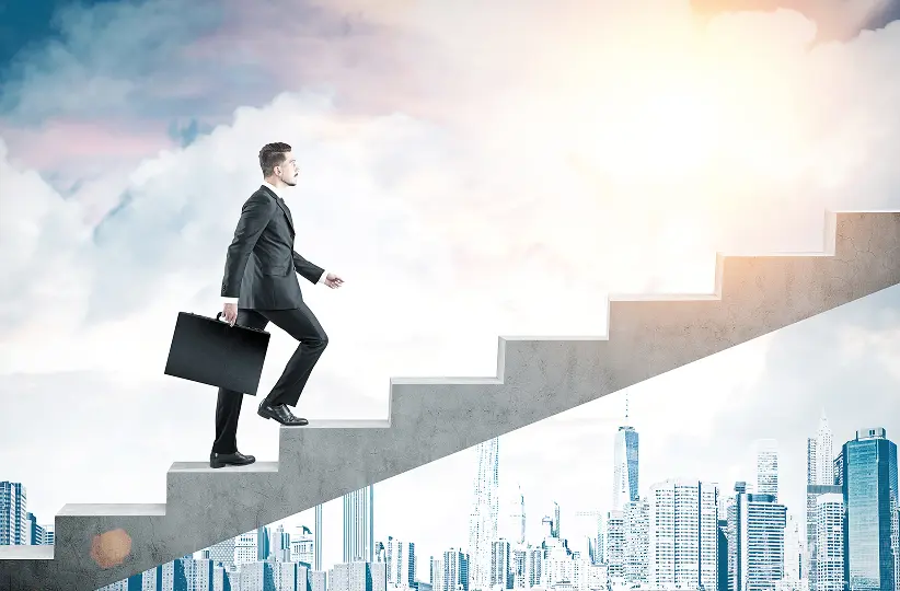 Businessman in suit carrying briefcase walking up concrete stairs above cityscape toward bright light.