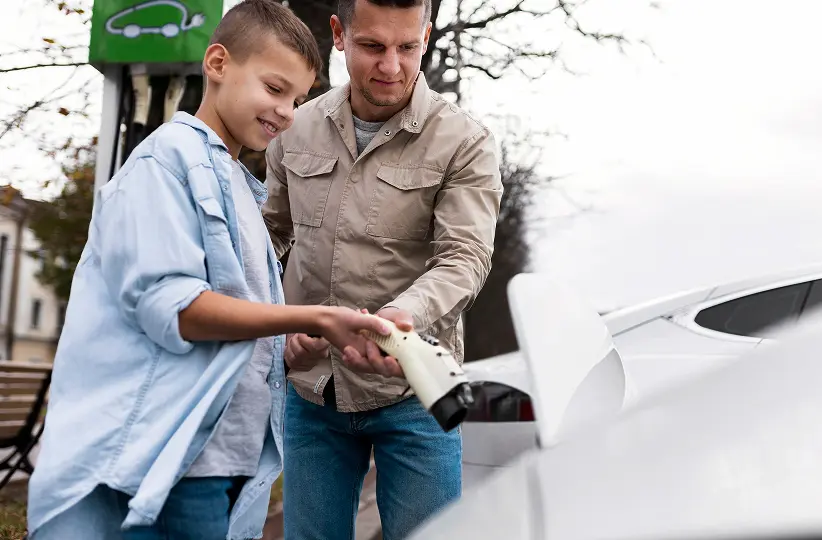 Man and boy connecting an electric car charging plug to a white vehicle outdoors.
