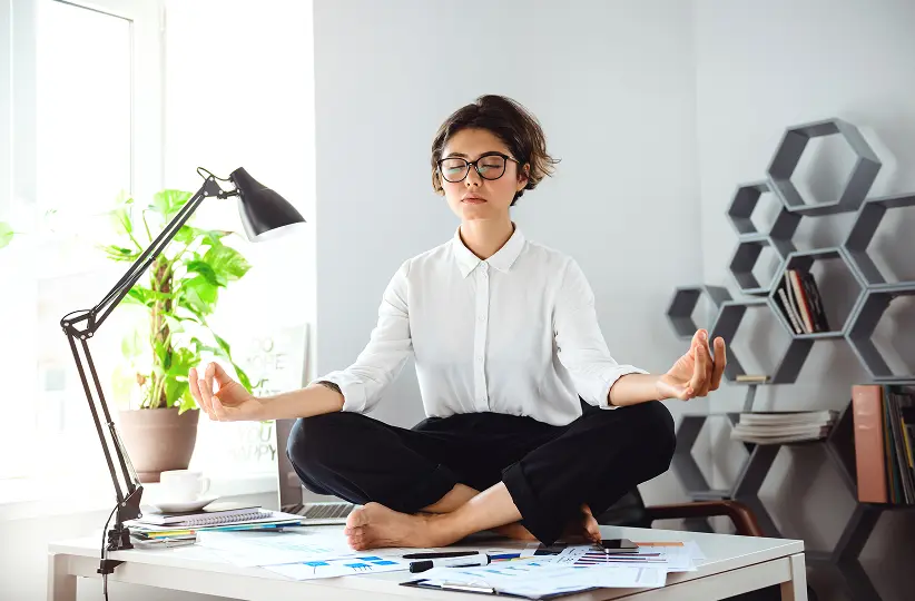 Woman in office attire meditating cross-legged on a desk covered with papers and stationery.
