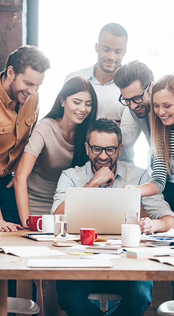 Smiling diverse group of coworkers gathered around a laptop at a wooden desk with mugs and papers.