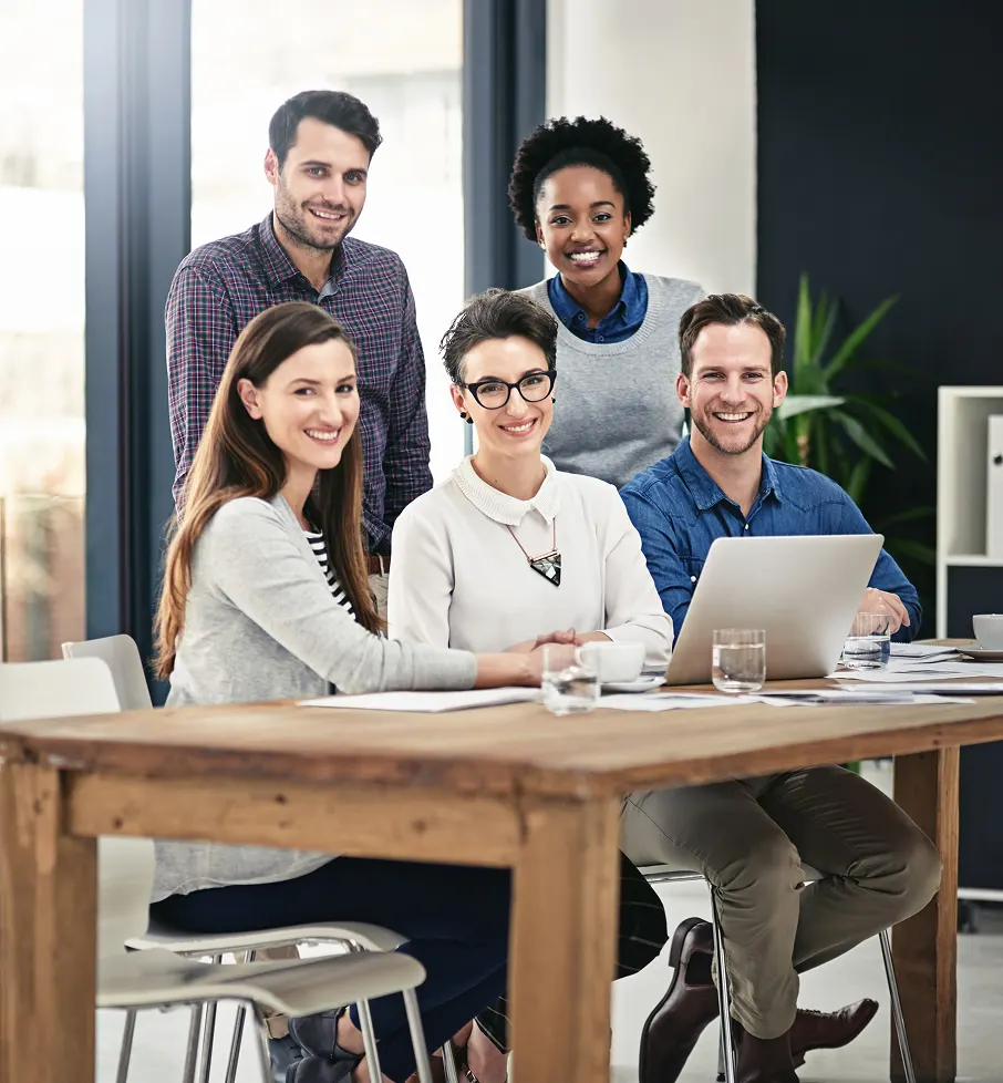 Group of five diverse professionals smiling around a wooden table with a laptop in a modern office.