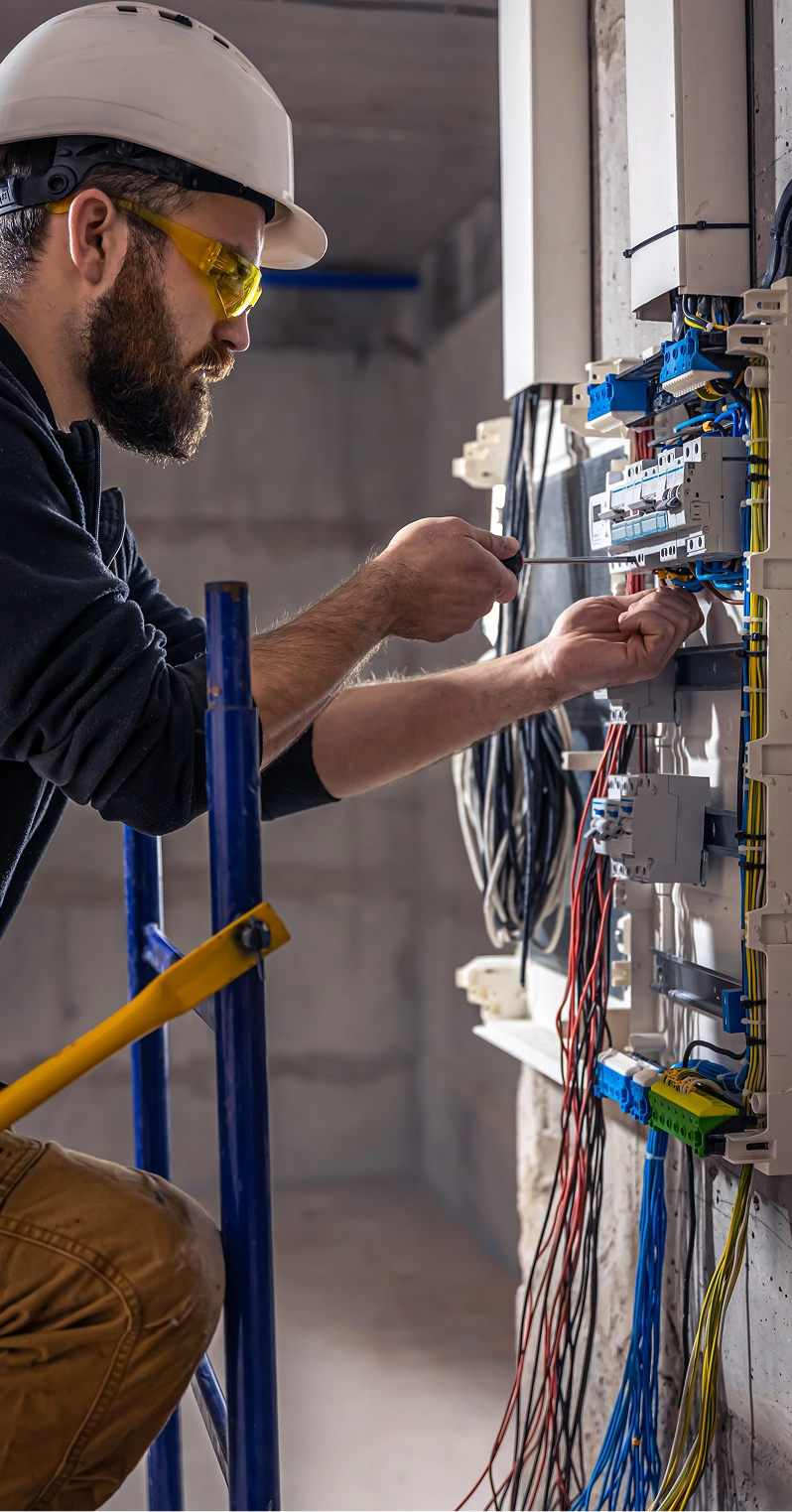 Electrician in safety helmet and glasses working with wiring inside an electrical panel.