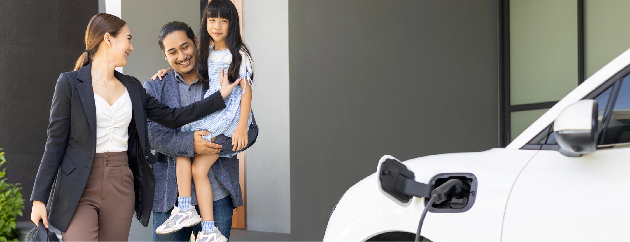 Family of three with a man holding a girl and a woman smiling near an electric car charging at home.