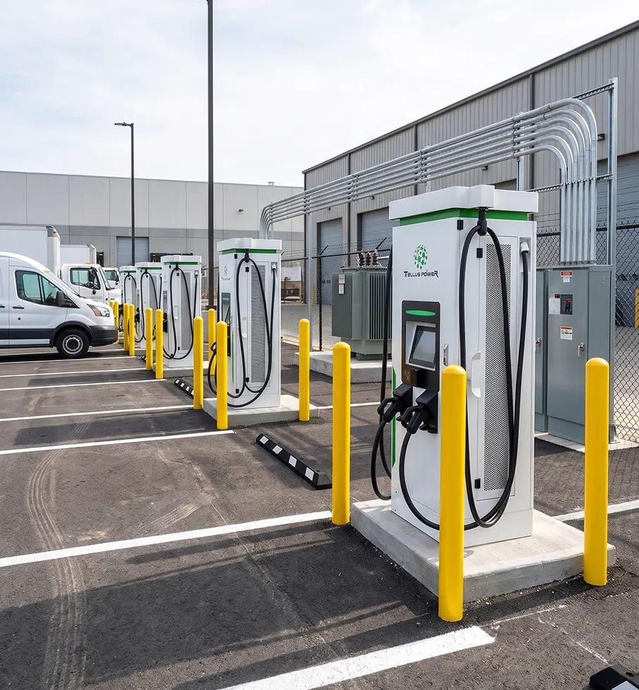 Row of electric vehicle charging stations with white vans parked and a fenced transformer area in a parking lot.