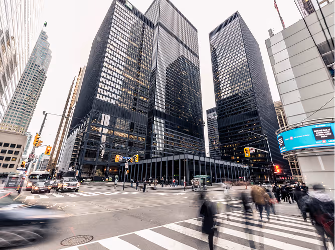 Busy urban street corner with pedestrians crossing and tall glass office buildings in the background.