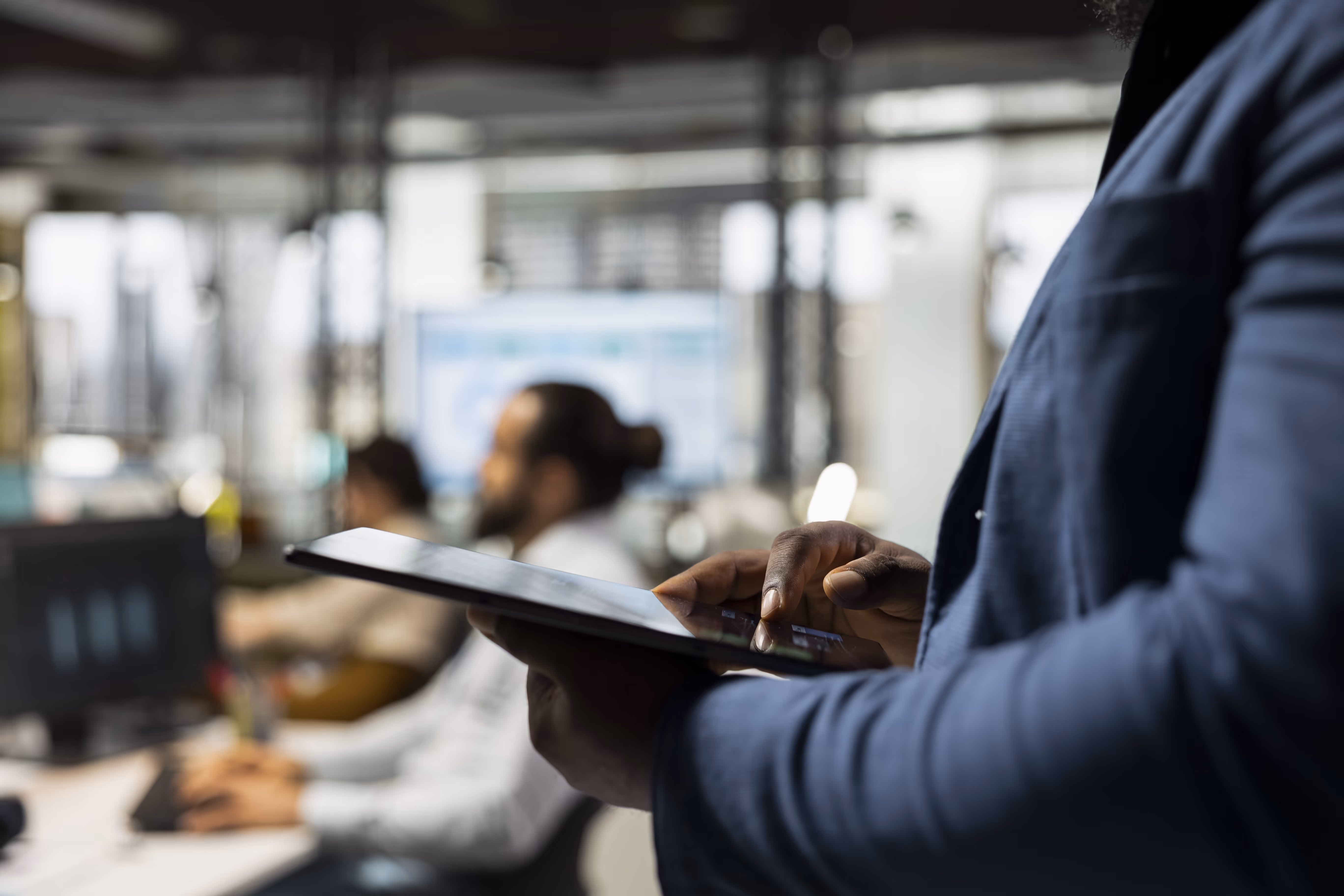 Close-up of a person using a tablet with blurred coworkers and monitors in the background.