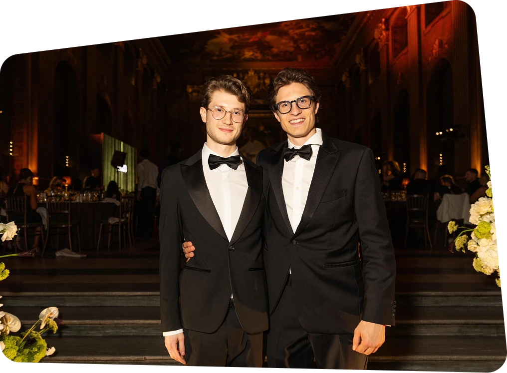 Two young men in black tuxedos and bow ties smiling at a formal event inside an ornate hall.