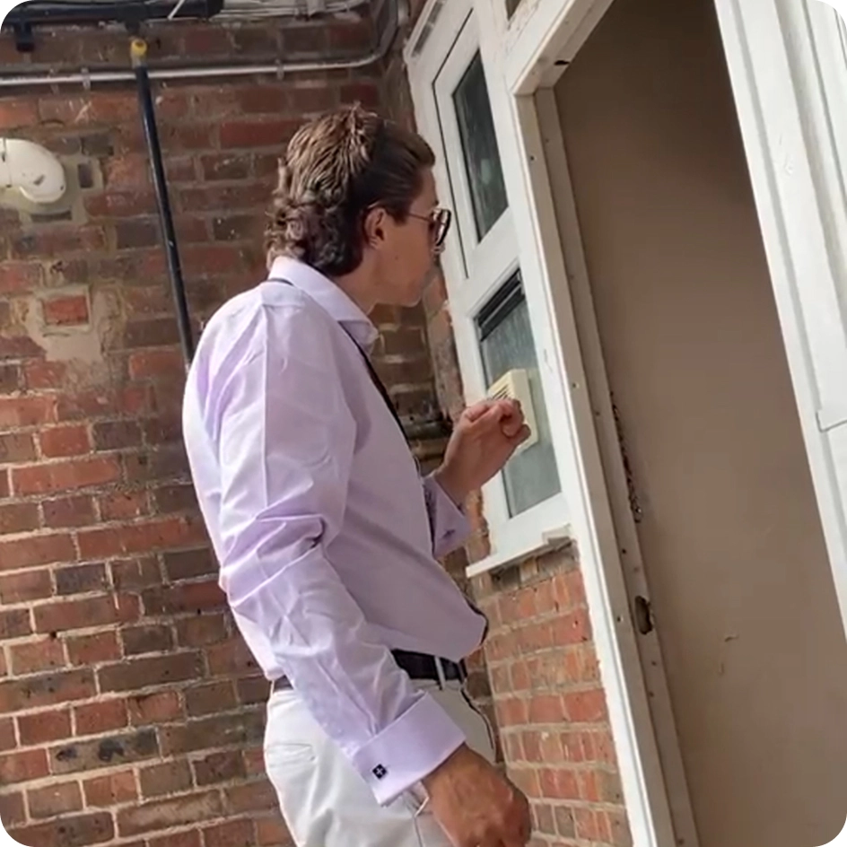 Man in glasses, light shirt, and light pants pressing a doorbell on a brick wall near a white door.
