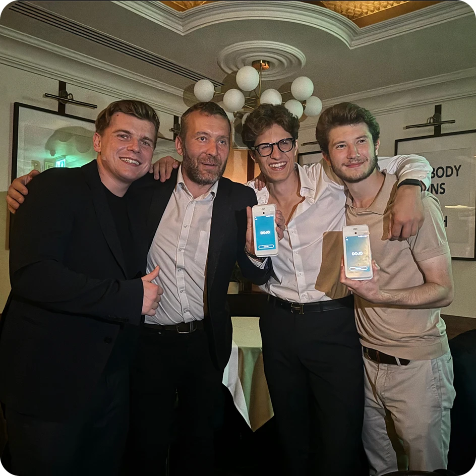 Four men smiling with arms around each other, two holding phones showing a blue app screen.