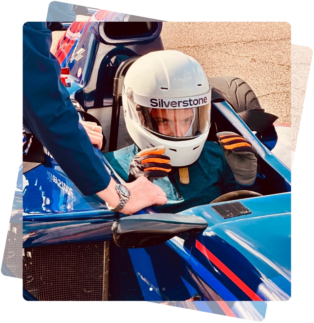 Race car driver in white Silverstone helmet inside blue open-wheel race car adjusting gloves with help from another person.