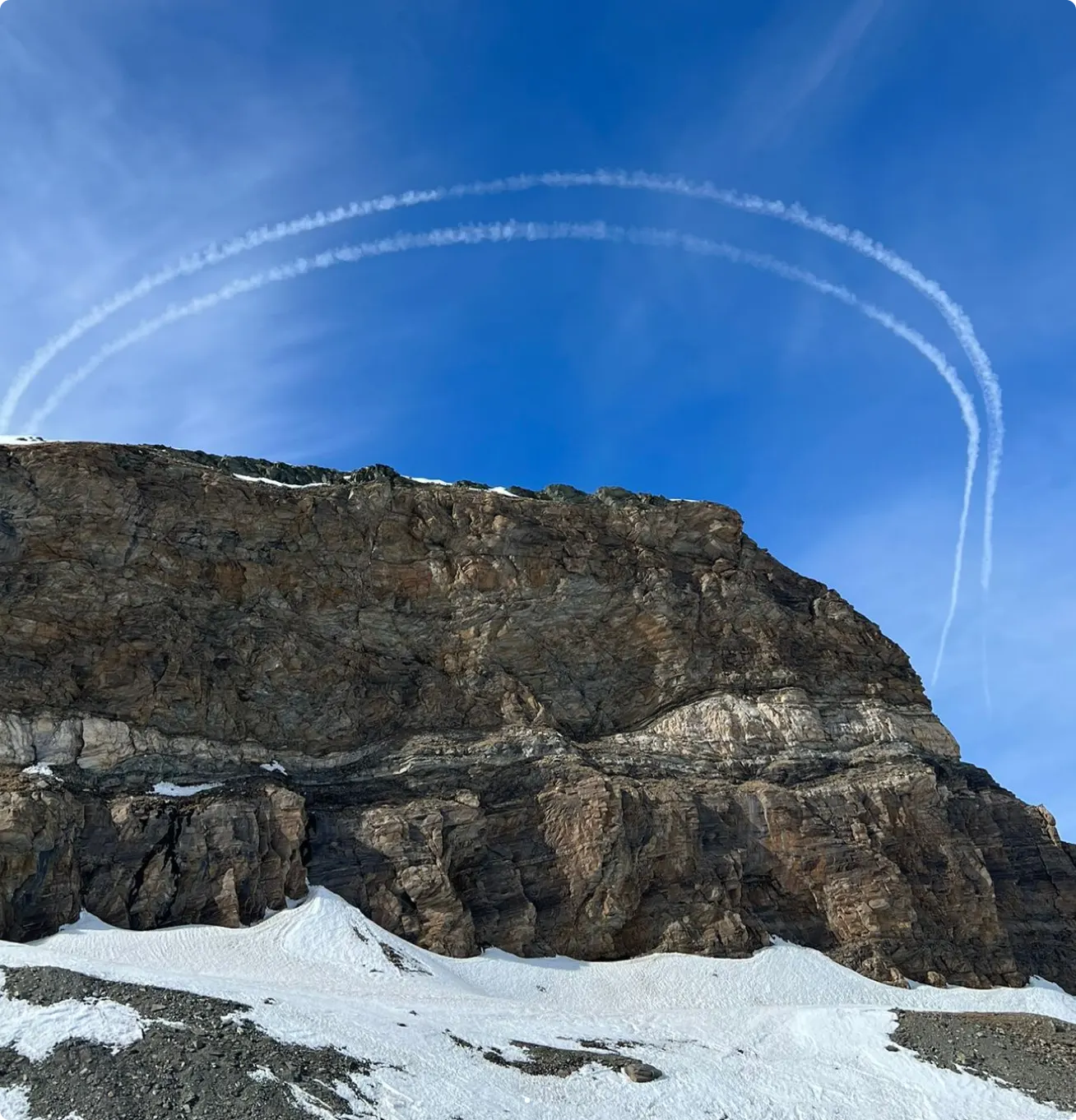 Rocky mountain cliff with snow at the base and twin curved contrails in a clear blue sky above.