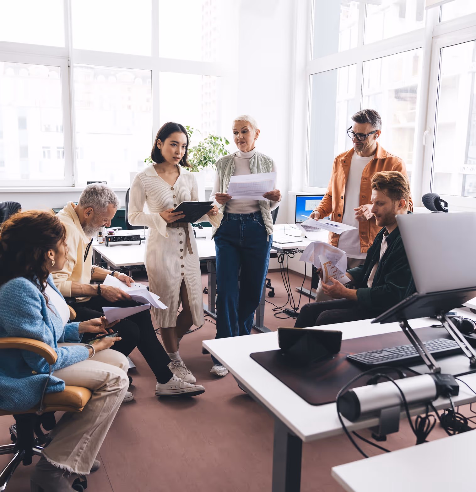 Six diverse coworkers in a bright office collaborating with documents and digital devices around desks.