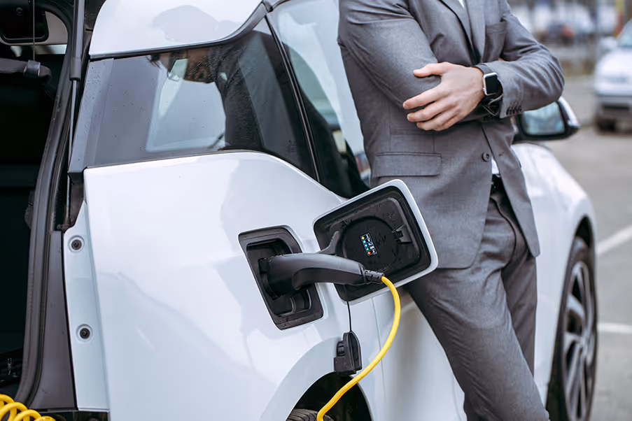 Person in a grey suit standing with arms crossed next to a white electric car that is charging with a plugged-in yellow cable.