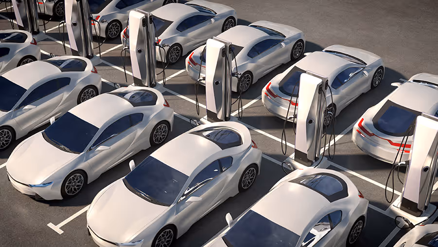 Rows of white electric cars parked and connected to charging stations in a parking lot.
