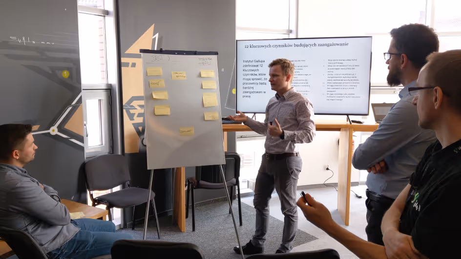 Man presenting ideas on a flip chart with sticky notes to a small group in a modern office space, with a screen showing a presentation in the background.