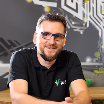 Smiling man with glasses and beard wearing a black i4e polo shirt sitting indoors with blurred tech-themed background.