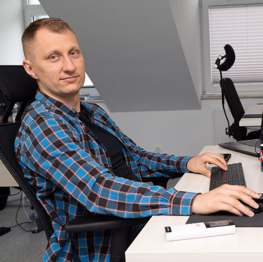 Man wearing a blue and orange plaid shirt sitting at a desk using a keyboard and mouse in an office.