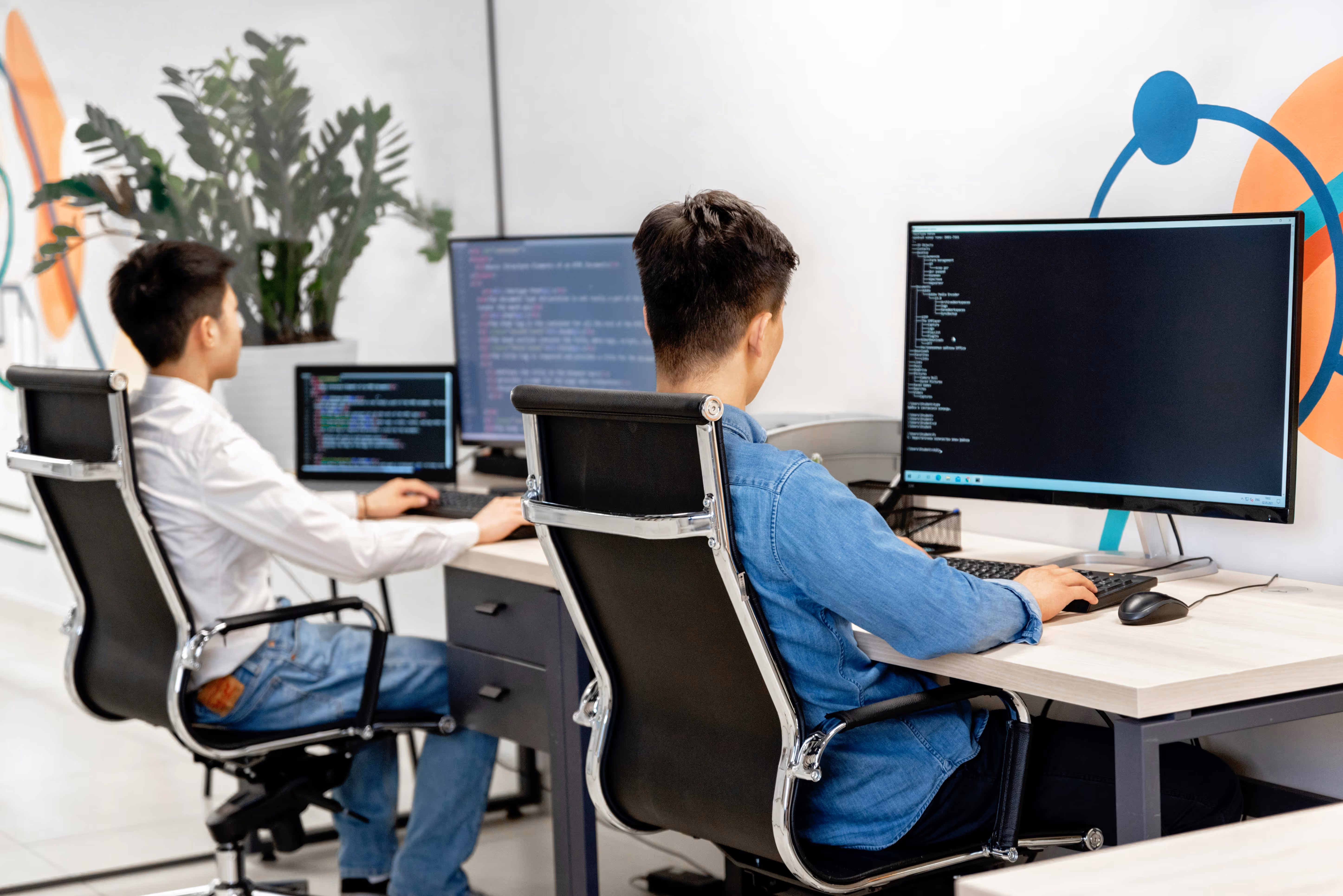 Two men seated at desks in an office, working on desktop computers with code displayed on their screens.