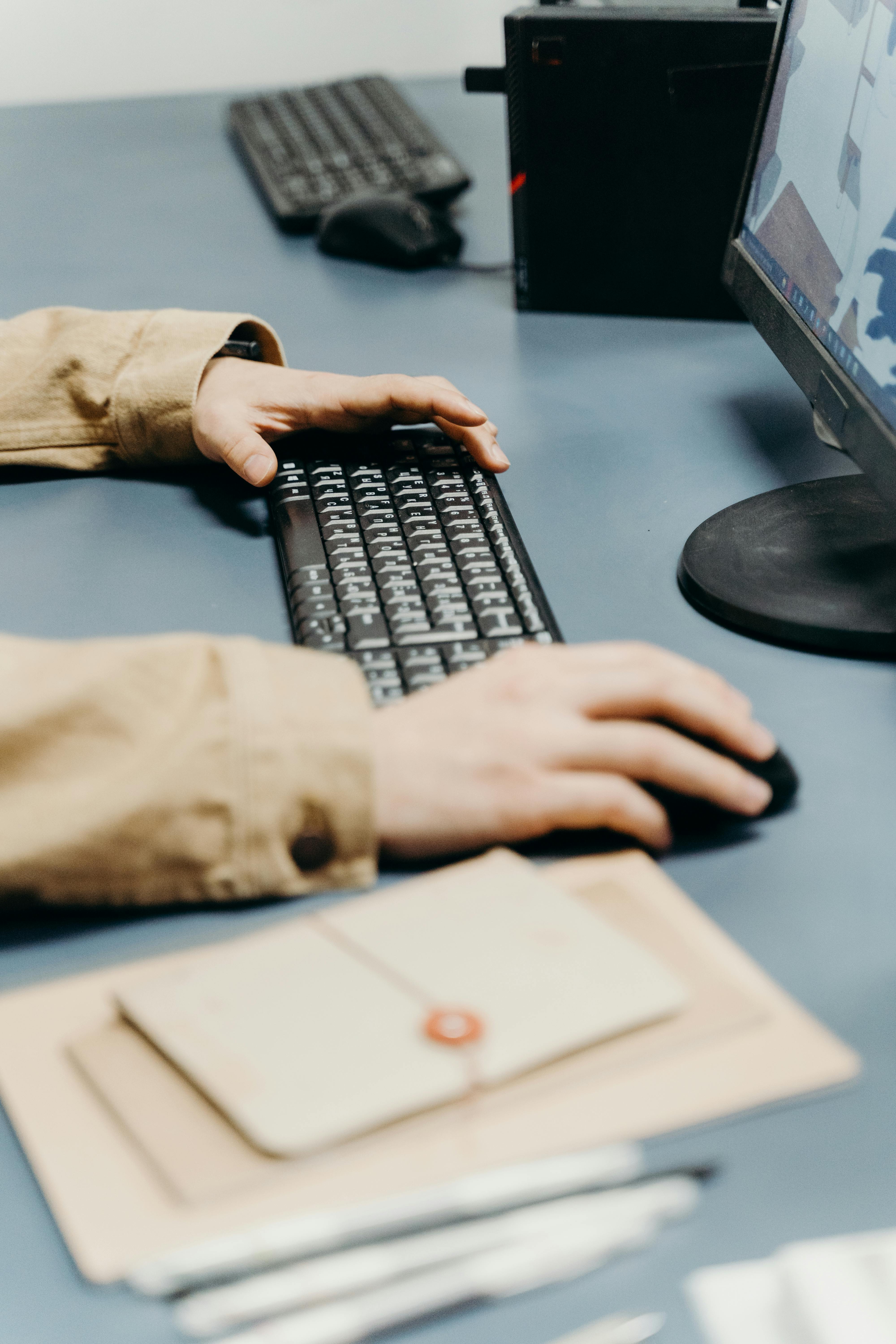 Person using a computer keyboard and mouse at a desk with a monitor, PC tower, and envelopes.