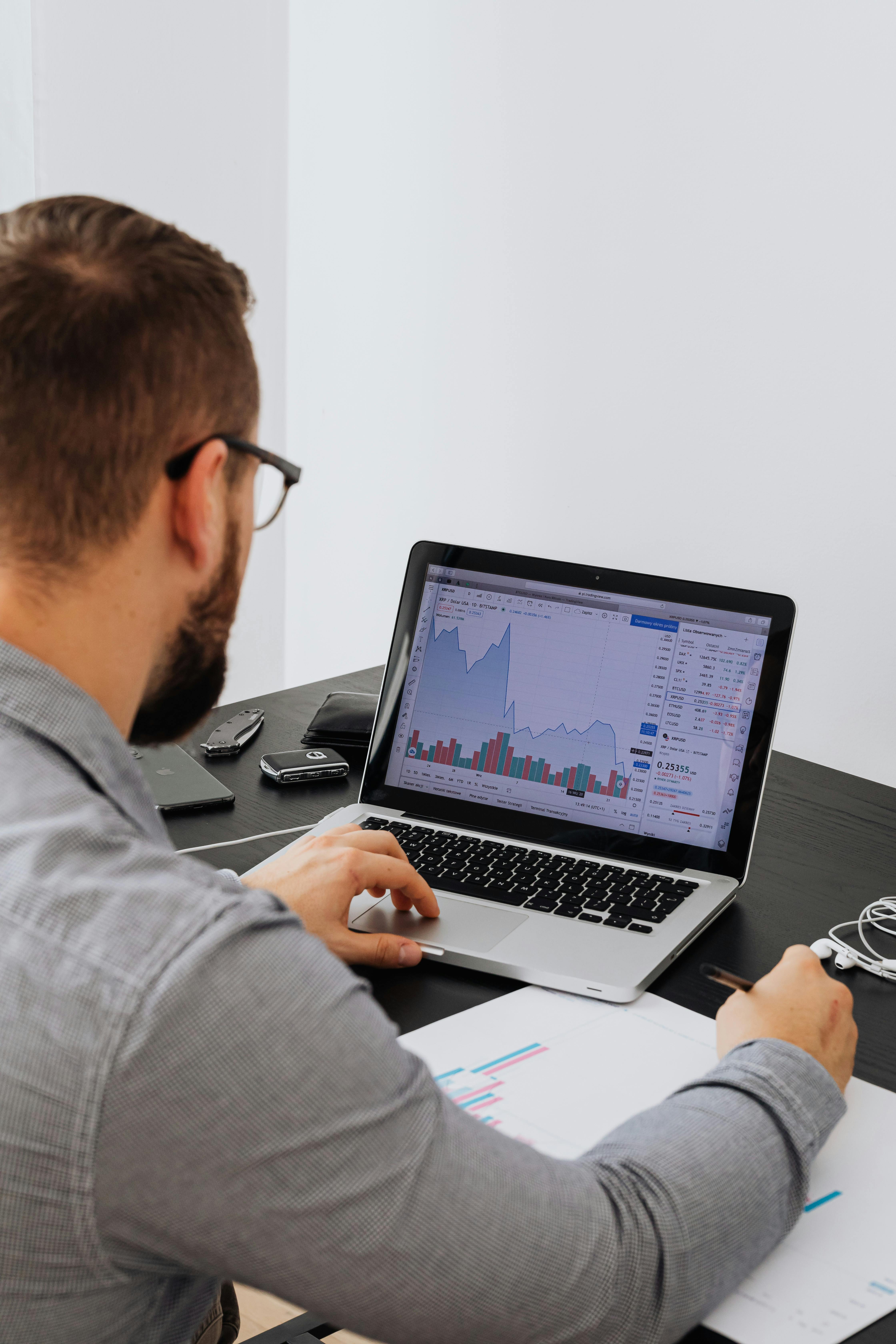 Man in glasses analyzing stock market data on a laptop with charts and graphs, holding a pen and paper with printed charts.