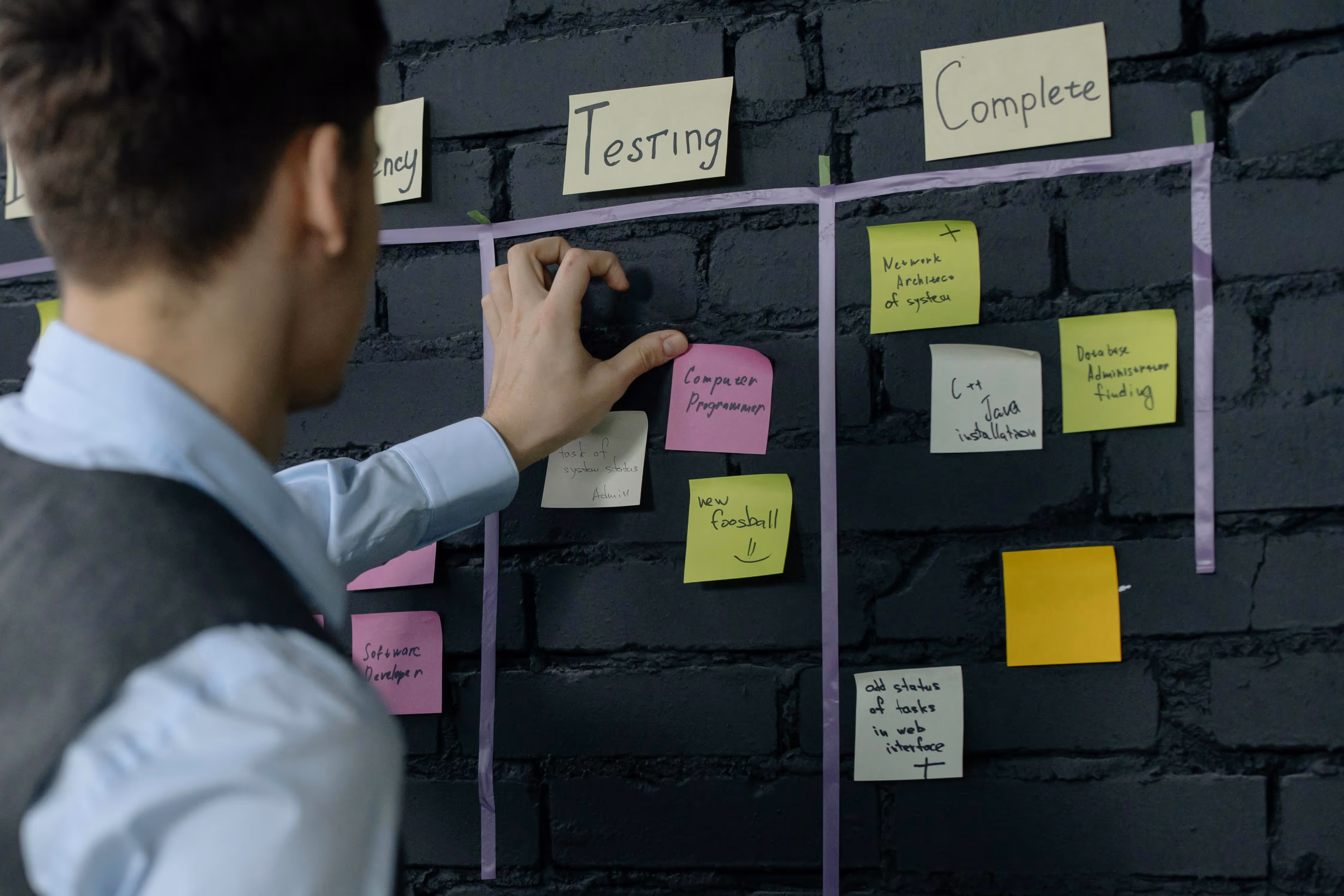 Man organizing colored sticky notes under labeled columns 'Testing' and 'Complete' on a black brick wall task board.