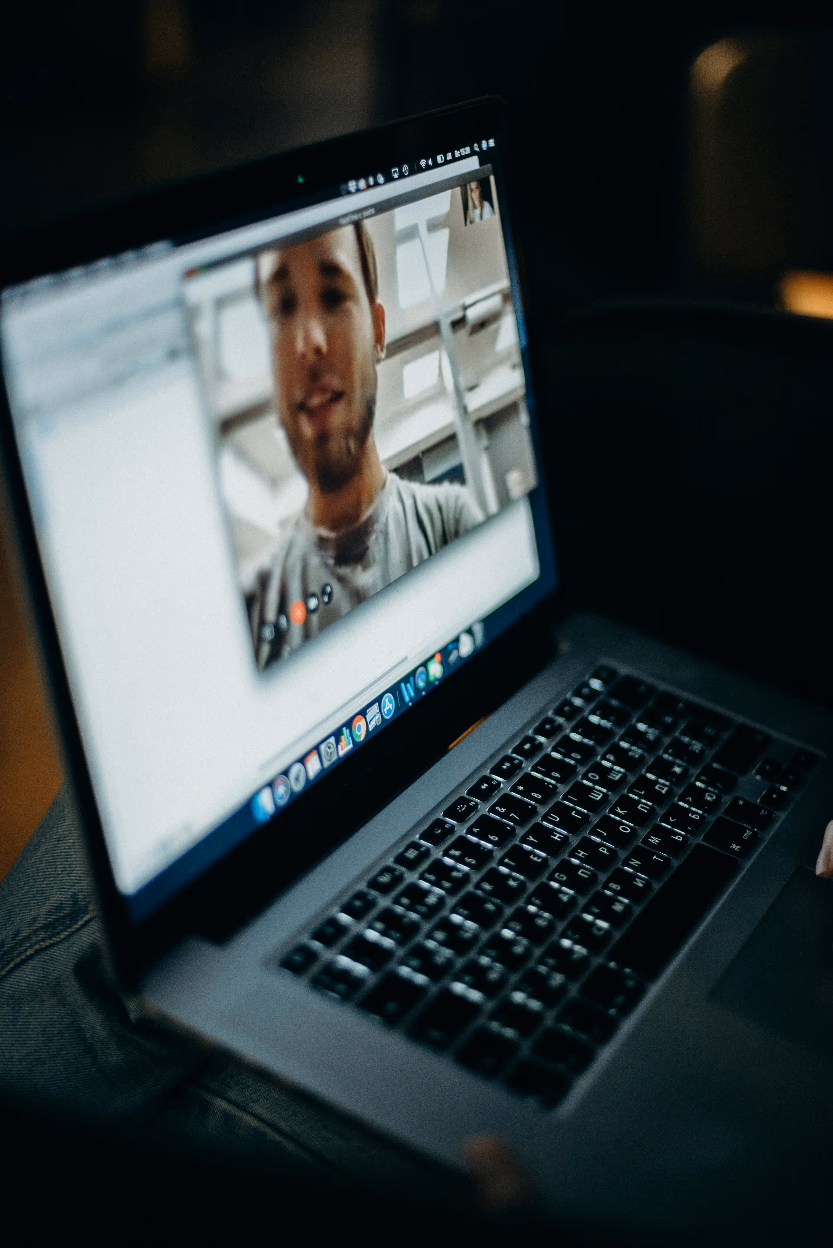 Laptop screen showing a video call with a man visible, keyboard and hand partly visible in low light.