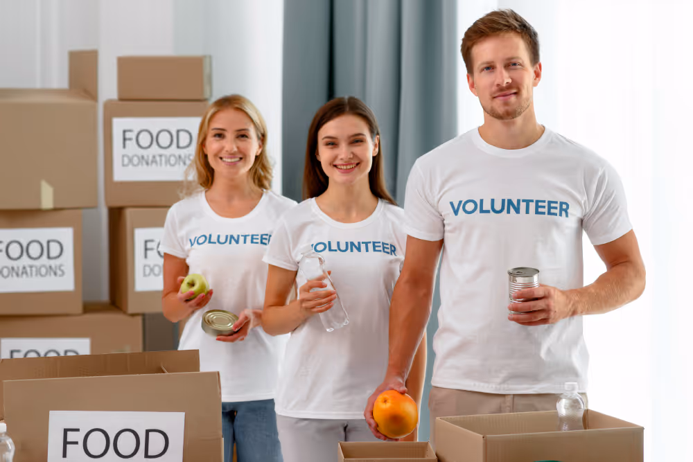 Three volunteers smiling, holding food items like an apple, a can, and a bottle, standing in front of boxes labeled 'FOOD DONATIONS'.