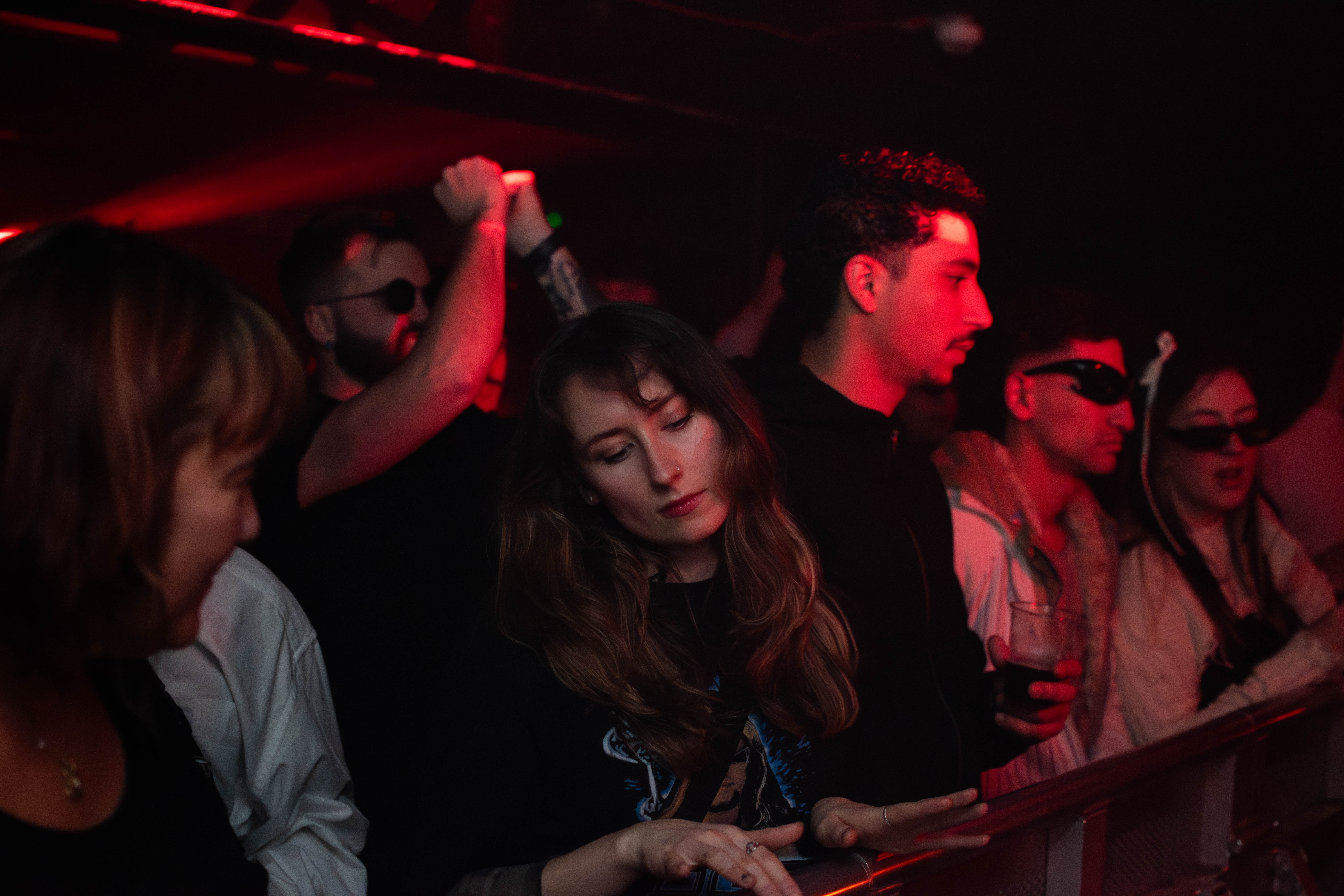 People standing close together in a dimly lit club with red lighting, some are dancing or holding drinks.