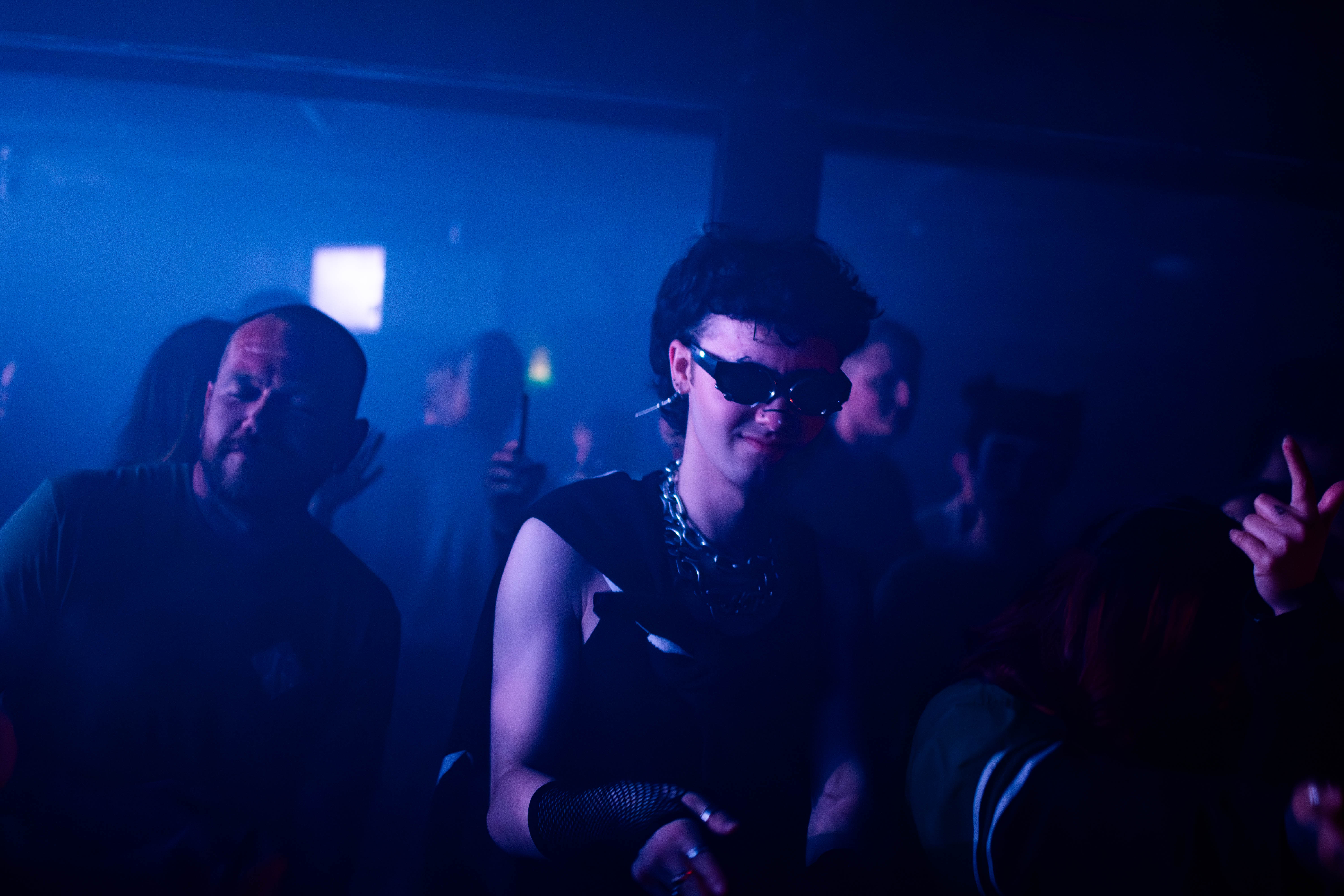 Person with dark sunglasses and chain necklace dancing in a dim, blue-lit club crowd.