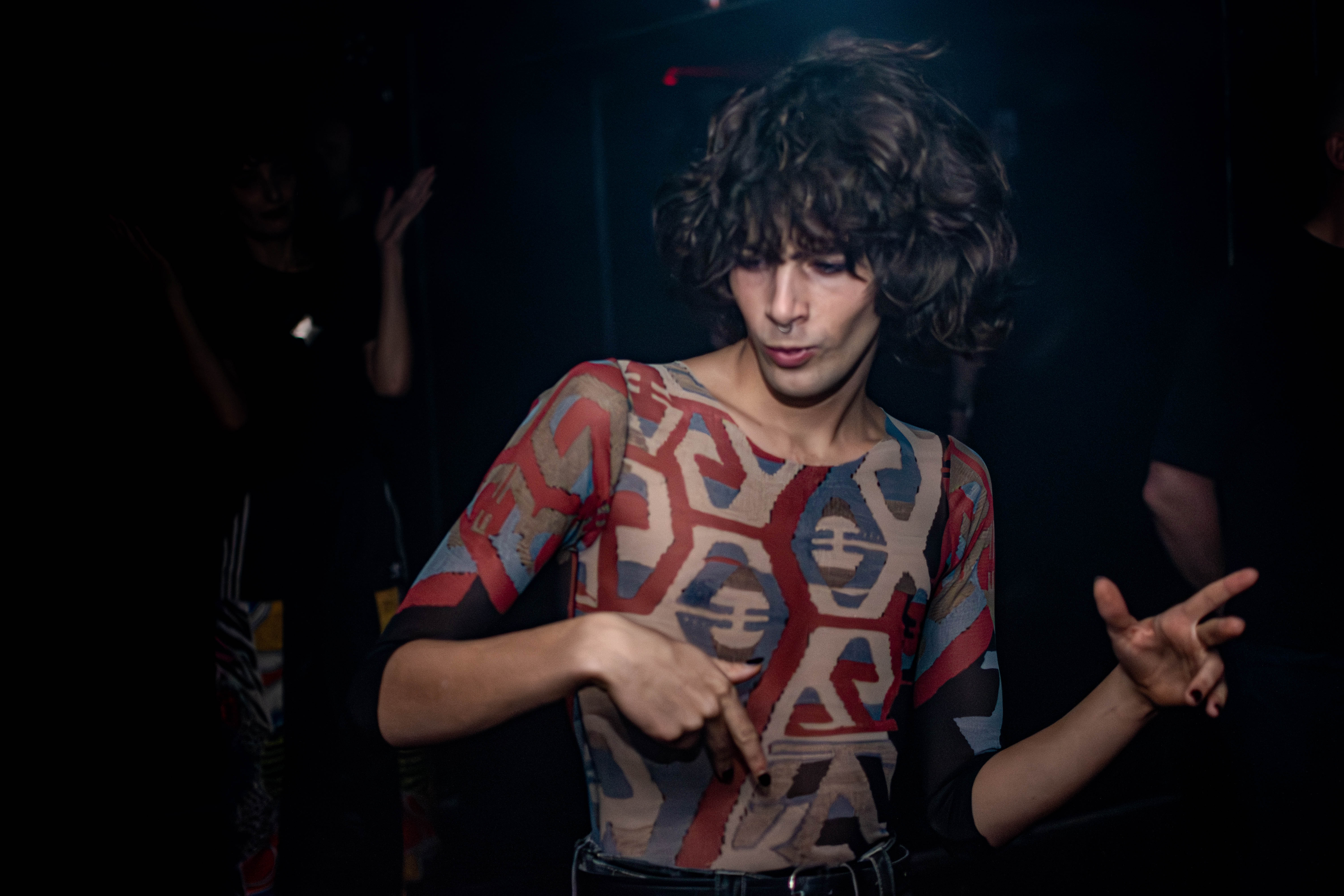 Person with curly hair wearing a patterned top dancing with hands pointing in different directions.