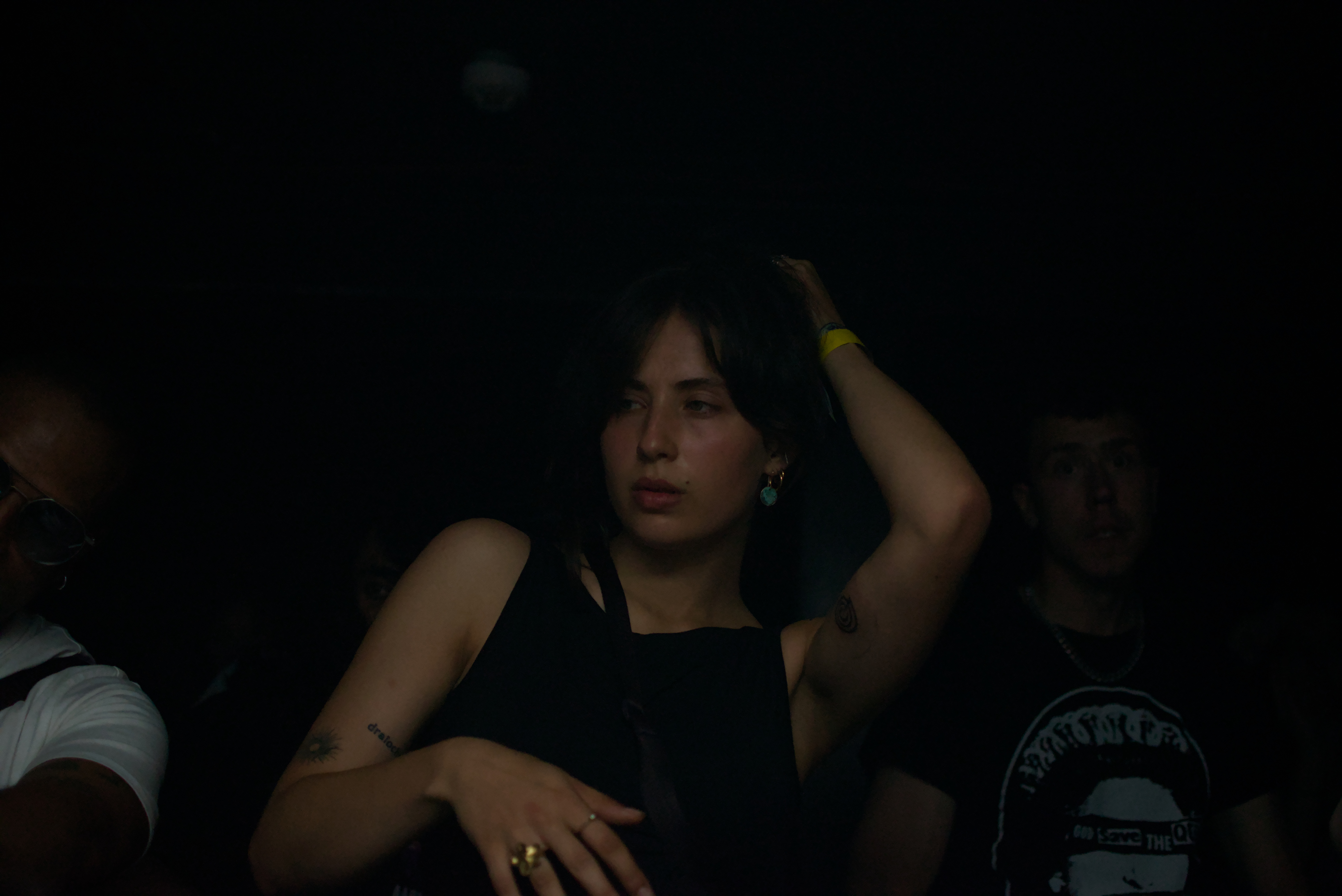 Young woman in a dark setting touching her hair wearing earrings and rings, with faint figures behind her.