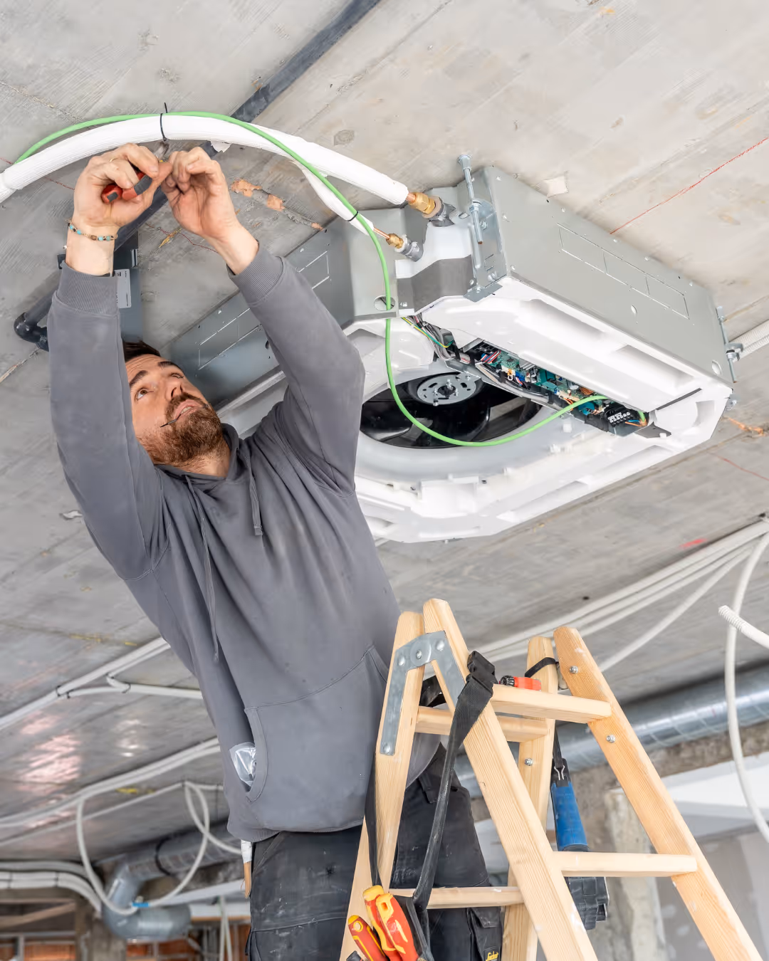 Technician standing on a ladder installing or repairing a ceiling-mounted HVAC unit with wires and pipes.