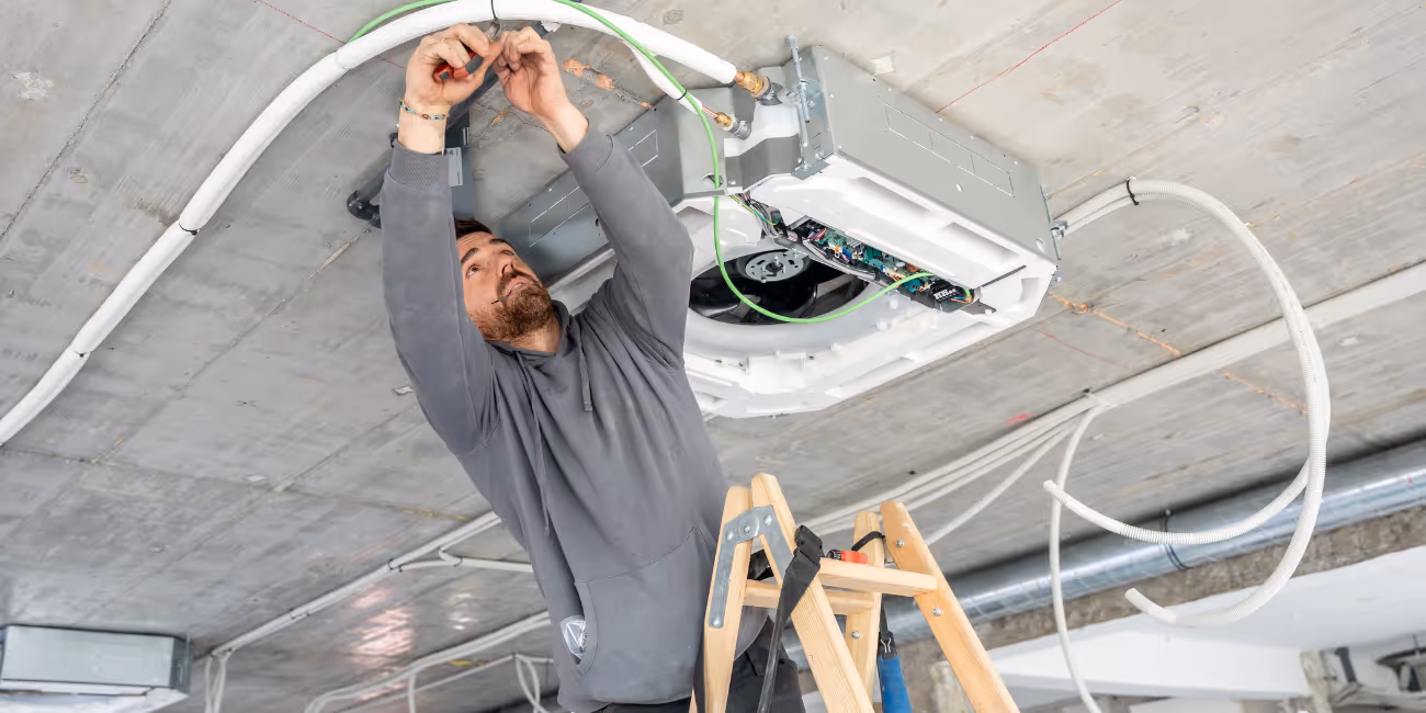Technician installing or repairing a ceiling-mounted HVAC unit while standing on a ladder.