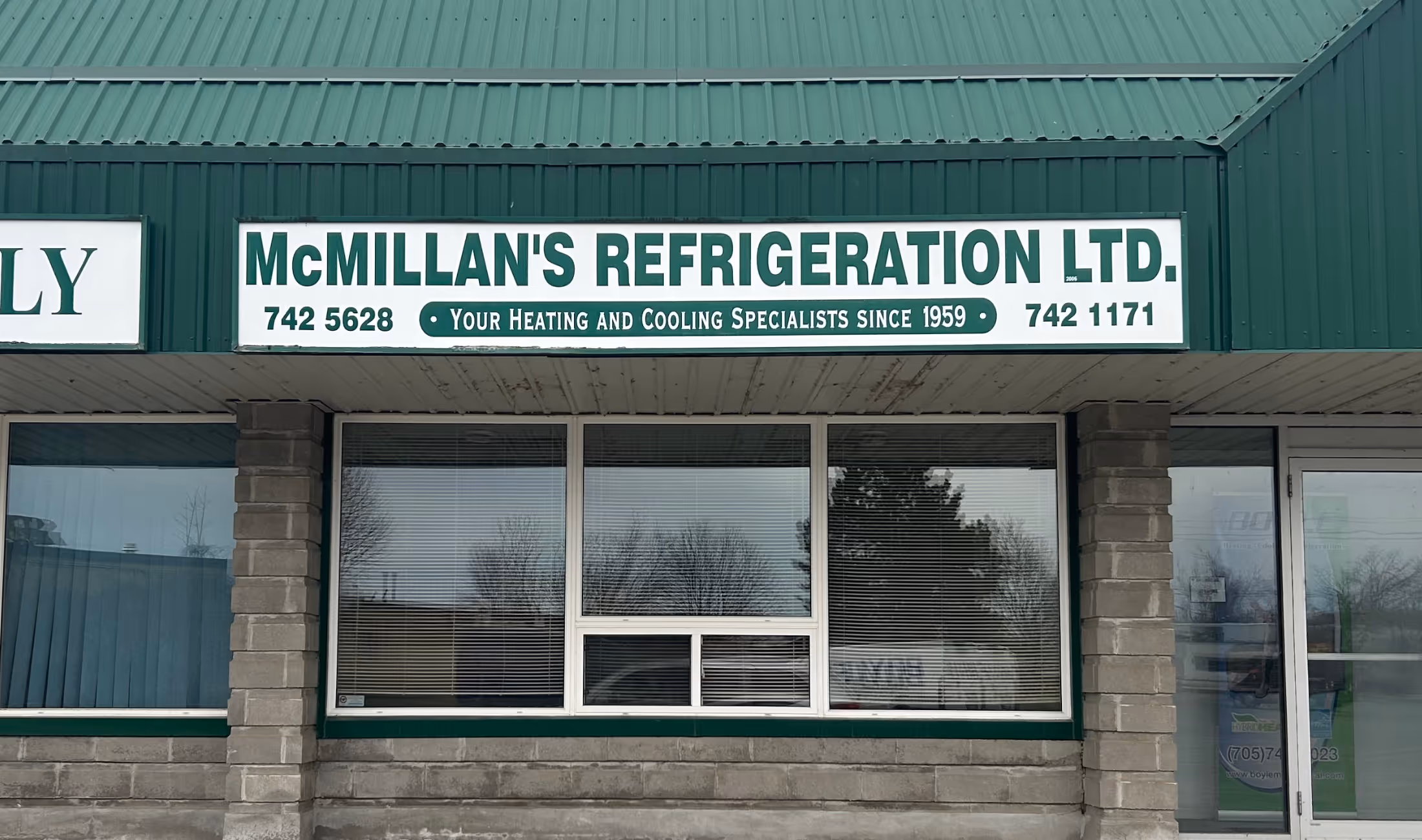 Storefront of McMillan's Refrigeration Ltd. with a green roof and sign displaying phone numbers and the tagline 'Your Heating and Cooling Specialists since 1959'.