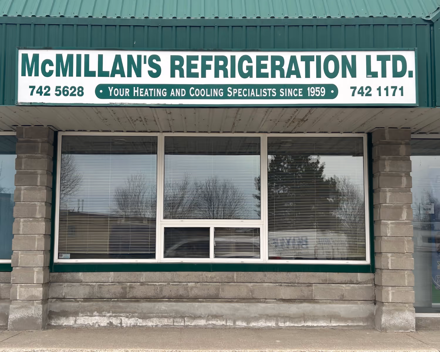 Front of McMillan's Refrigeration Ltd. building with a green and white sign displaying phone numbers and the slogan 'Your Heating and Cooling Specialists since 1959'.