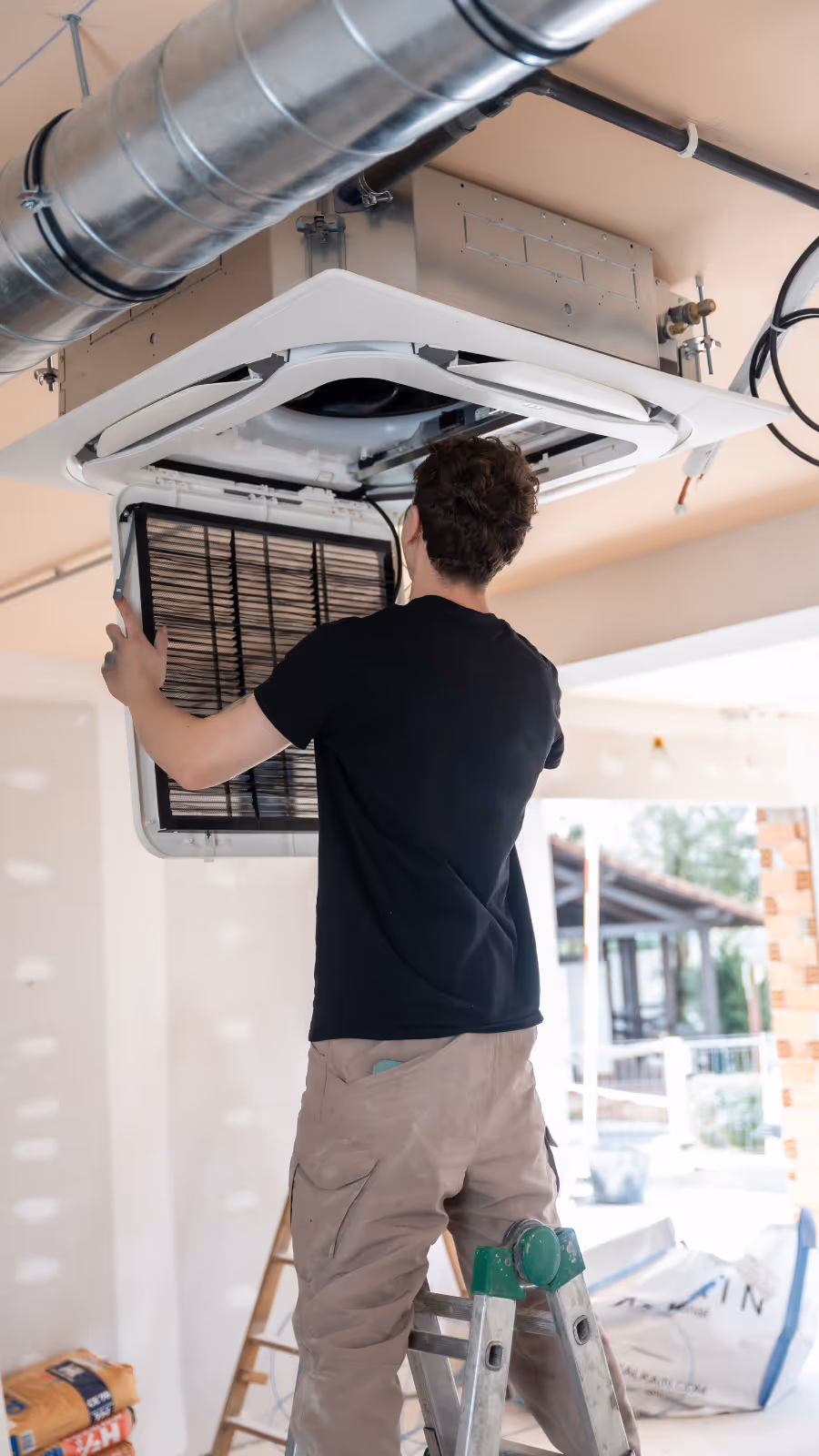 Technician standing on a ladder installing or repairing a ceiling-mounted HVAC air conditioning unit.