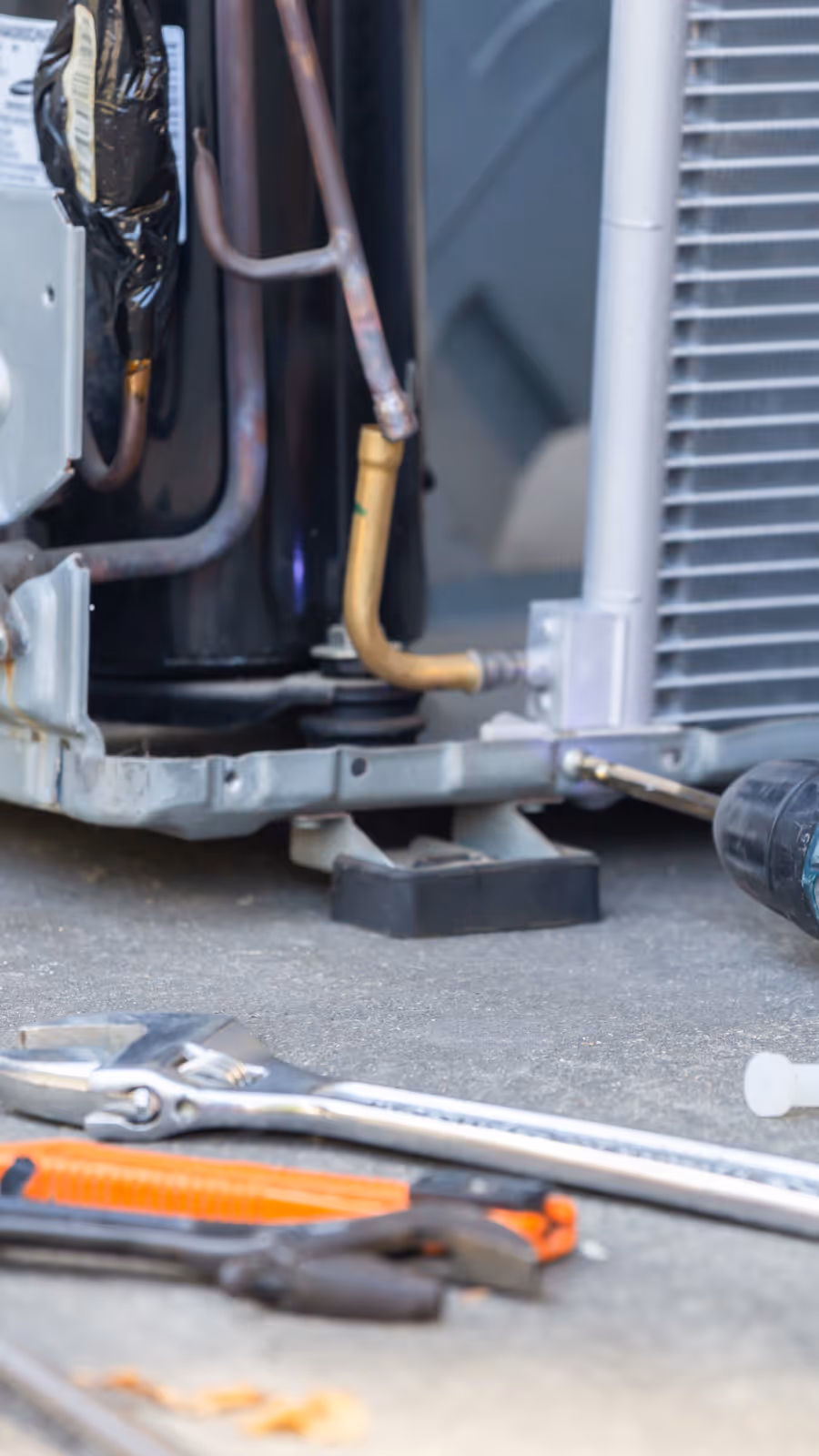 Close-up of HVAC unit components and tools including a wrench, pliers, and a drill on the ground.