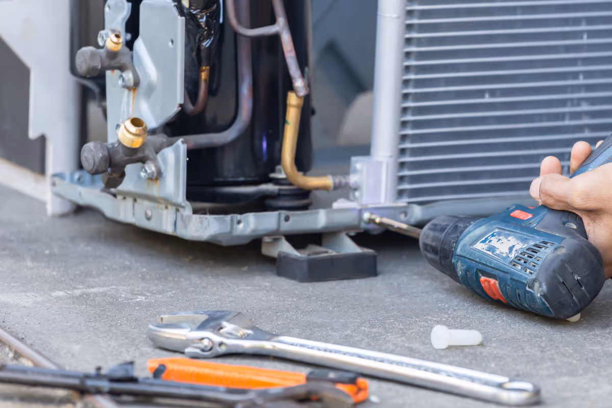 Hand holding a power drill tightening a screw on an air conditioning unit with a wrench and utility knife on the ground.