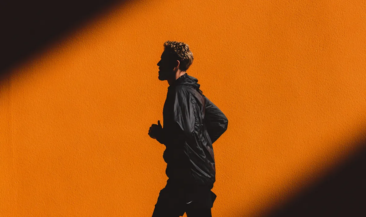 Silhouette of a man jogging in a black jacket against an orange wall with shadows.