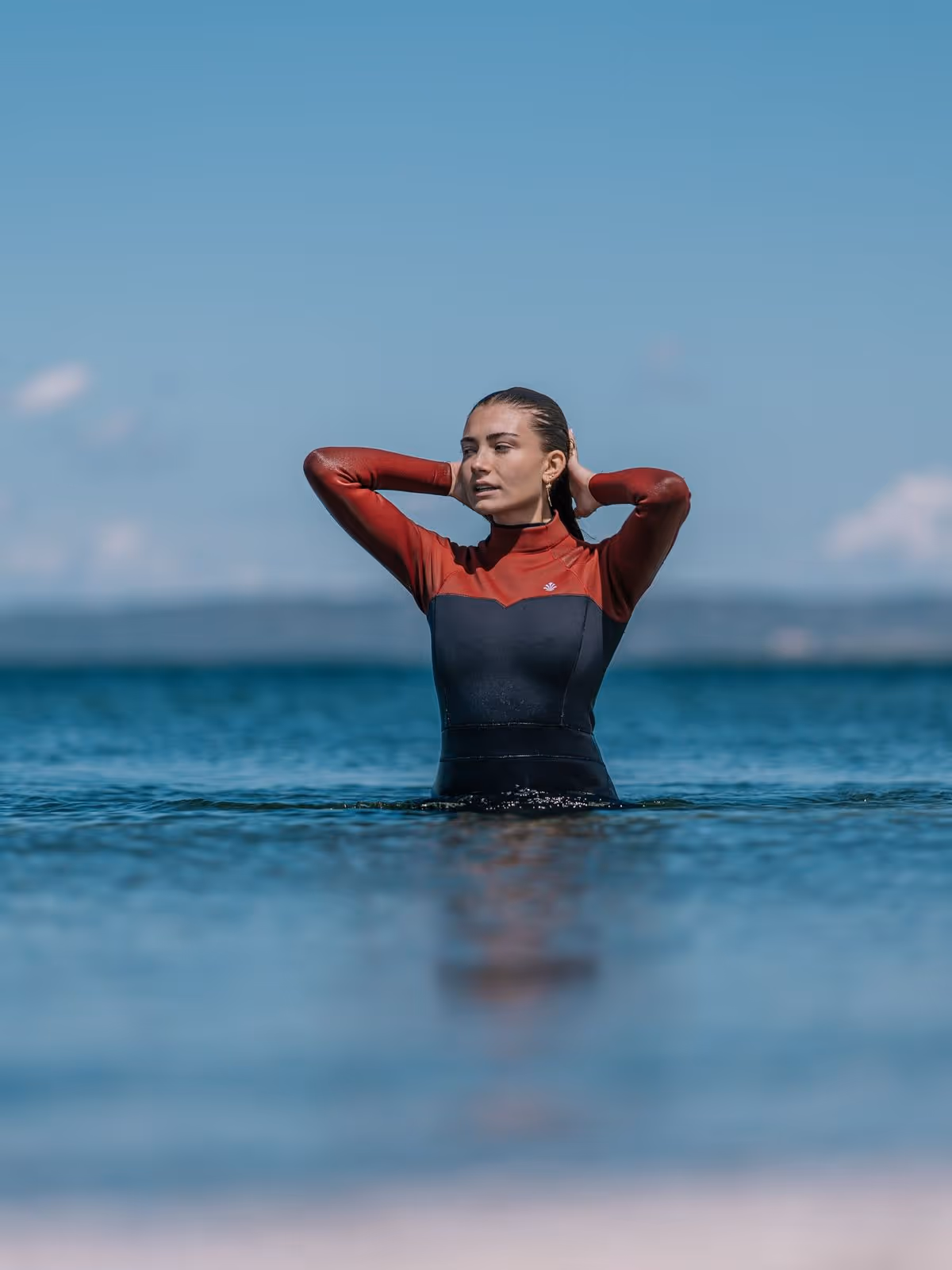 Woman in a red and black wetsuit standing waist-deep in calm water with hands behind her head.