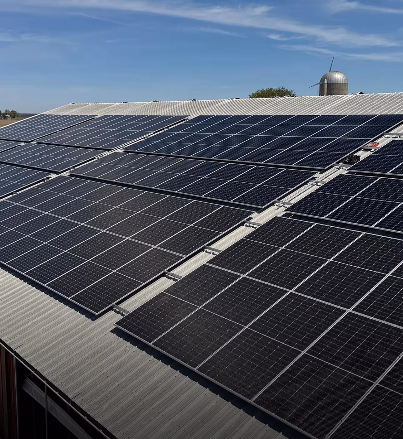 Rows of solar panels installed on a corrugated metal rooftop under a blue sky with a wind turbine in the background.