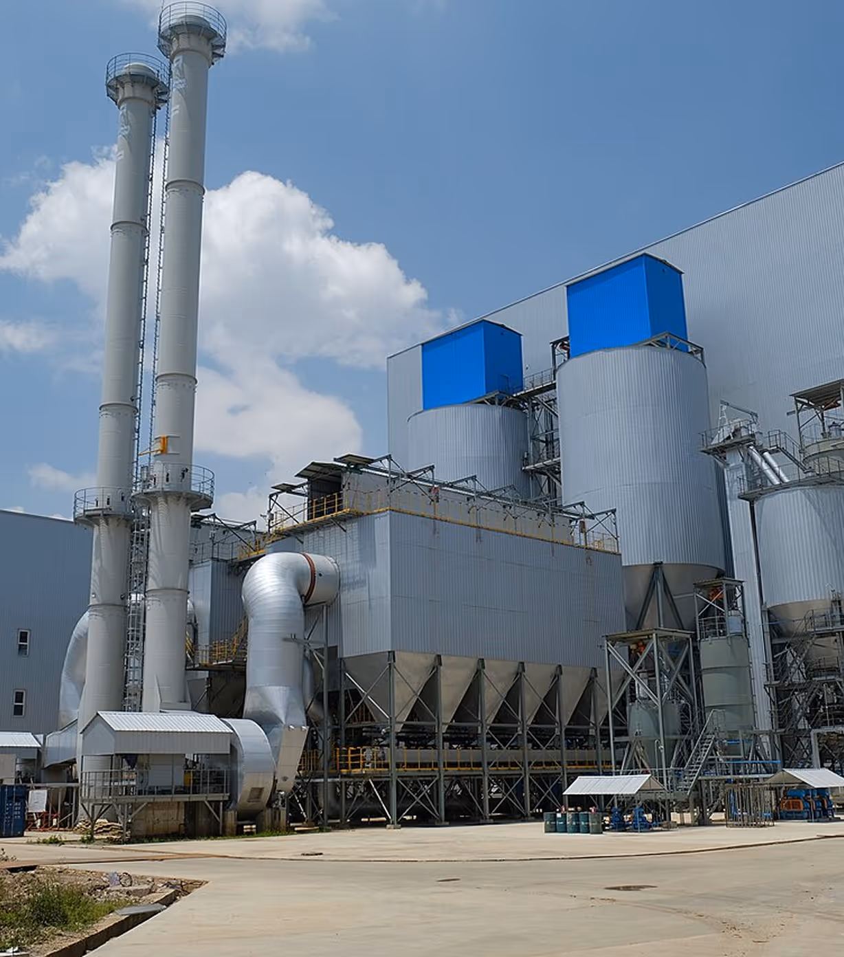 Industrial facility with tall chimneys, large cylindrical storage tanks with blue tops, and metallic ductwork under a partly cloudy blue sky.