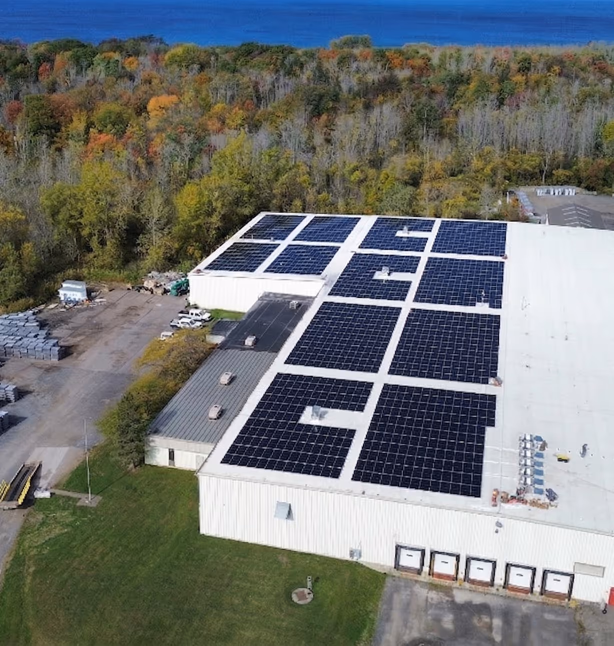 Aerial view of a large industrial building with multiple solar panel arrays installed on the roof, surrounded by green grassy areas and trees with autumn foliage in the background.